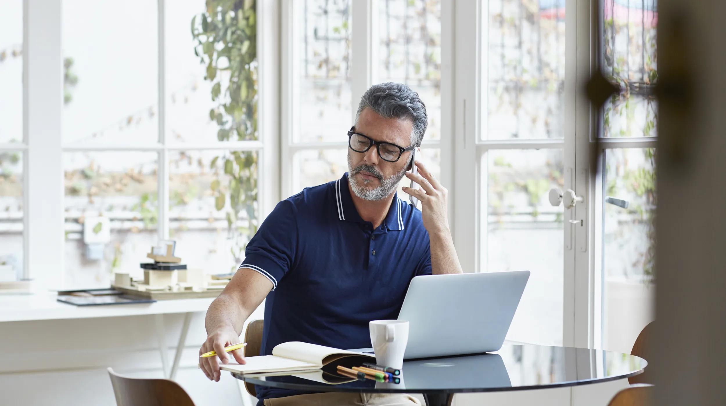Man working at a table with a laptop, notebook, and coffee, talking on the phone.