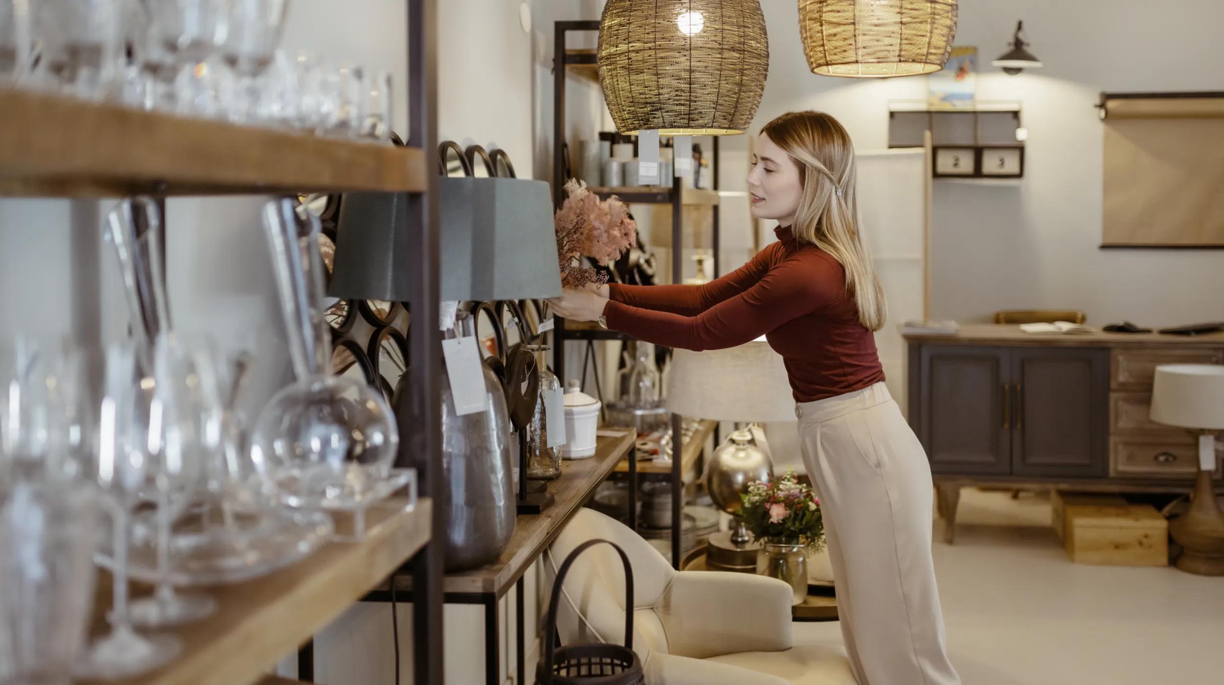 A woman organizing decorative items on a shelf in a home goods store, surrounded by glassware and lamps, representing a retail business environment where having a wholesale license is important for purchasing inventory at discounted prices.