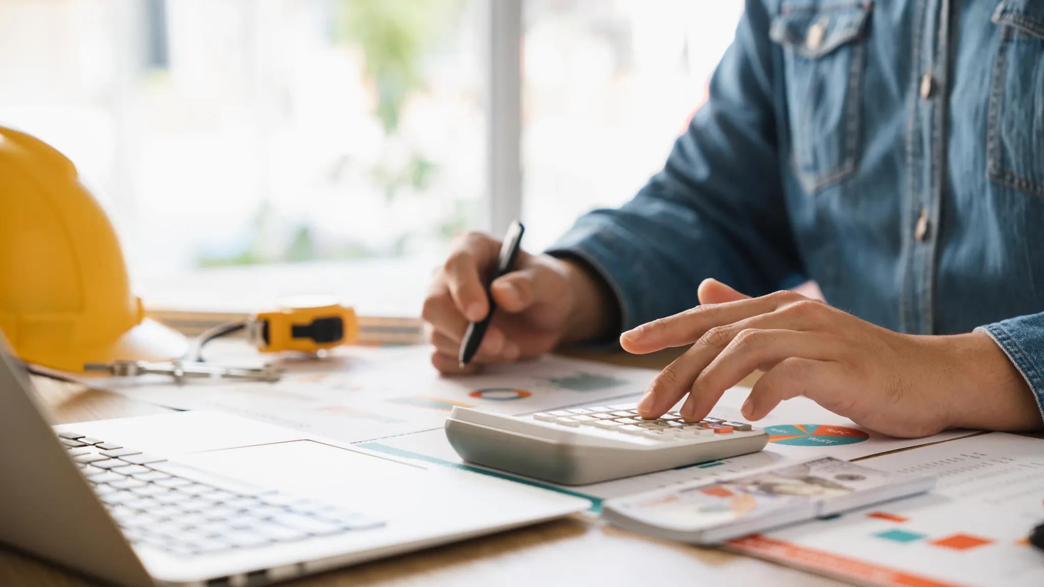 Person working with business documents and a calculator with a hard hat resting on the desk beside them.