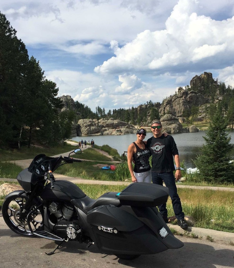 a man and woman standing next to a motorcycle in front of a lake